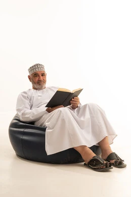 تعابير التمعن والتركيز، ممارسة هواية القراءة, قضاء وقت الفراغ بمطالعة الكتب والروايات, بورتريه لرجل مسن عربي خليجي عماني يرتدي الزي التقليدي يجلس على كرسي مريح، خلفية بيضاء