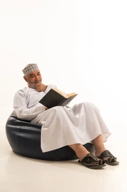 تعابير التمعن والتركيز، ممارسة هواية القراءة, قضاء وقت الفراغ بمطالعة الكتب والروايات, بورتريه لرجل مسن عربي خليجي عماني يرتدي الزي التقليدي يجلس على كرسي مريح، خلفية بيضاء