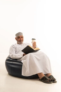 Practicing the hobby of reading, spending free time sipping a favorite drink, sitting and enjoying hot beverages and warm winter herbs, a portrait of an elderly Arab Gulf Omani man wearing traditional attire sipping Arabic coffee, white background. Practicing the hobby of reading, spending free time sipping a favorite drink, sitting and enjoying hot beverages and warm winter herbs, a portrait of an elderly Arab Gulf Omani man wearing traditional attire sipping Arabic coffee, white background.