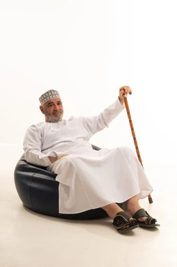 The need to use a cane, elegance and handsomeness and attention to external appearance, gestures of seriousness and self-confidence, a close-up portrait of an elderly Omani Gulf Arab man wearing traditional attire sitting on a comfortable chair holding a cane in his hand, white background.
