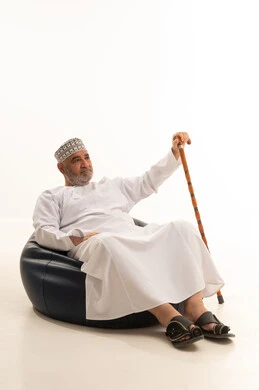 الحاجة إلى الاستعانة بعكاز، الأناقة والوسامة والاهتمام بالمظهر الخارجي، إيماءات الجدية والثقة بالنفس، بورتريه مقرب لرجل عربي خليجي عماني كبير السن يرتدي الزي التقليدي يجلس على كرسي مريح يمسك بيده عكاز، خلفية بيضاء