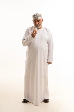 النظر الى الكاميرا بإيماءات السرور والسعادة, قضاء وقت الفراغ باحتساء المشروب المفضل, الجلوس والاستمتاع بالمشروبات الساخنة والأعشاب الشتوية الدافئة, بورتريه لرجل مسن عربي خليجي عماني يرتدي الزي التقليدي يقوم باحتساء القهوة العربية , الجسم بالطول الكامل، خلفية بيضاء