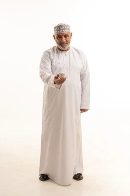 Looking at the camera with gestures of joy and happiness, spending leisure time sipping a favorite drink, sitting and enjoying hot beverages and warm winter herbs, a portrait of an elderly Arab Gulf Omani man wearing traditional attire sipping Arabic coffee, full body, white background.