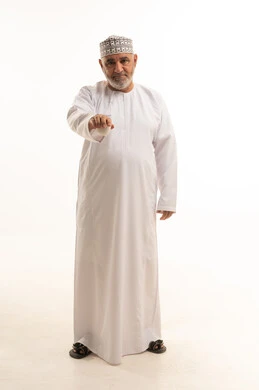 النظر الى الكاميرا بإيماءات السرور والسعادة, قضاء وقت الفراغ باحتساء المشروب المفضل, الجلوس والاستمتاع بالمشروبات الساخنة والأعشاب الشتوية الدافئة, بورتريه لرجل مسن عربي خليجي عماني يرتدي الزي التقليدي يقوم باحتساء القهوة العربية , الجسم بالطول الكامل، خلفية بيضاء