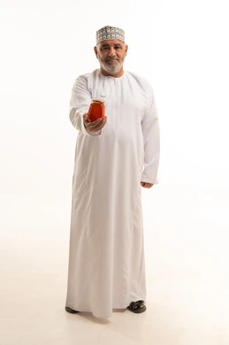 Looking at the camera with gestures of joy and happiness, a portrait of an elderly Omani Gulf Arab man wearing traditional attire holding a glass jar of fresh honey in his hand, local Saudi national products, healthy natural food, full-body shot, white background.