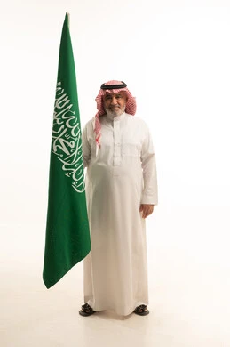 الفخر و الاعتزاز بالانتماء للمملكة, بورتريه لرجل مسن عربي سعودي خليجي مبتسم يرتدي الشماغ والثوب التقليدي يحمل بيده علم السعودية , يوم العلم 11مارس، الاحتفال بذكرى اليوم الوطني السعودي 8 ربيع الأول, الجسم بالطول الكامل، خلفية بيضاء