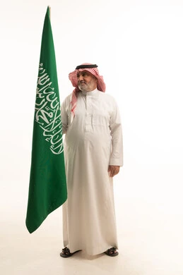 Pride and honor in belonging to the Kingdom, a portrait of an elderly Arab Saudi Gulf man smiling, wearing a ghutrah and traditional thobe, holding the Saudi flag in his hand, Flag Day March 11, celebrating the anniversary of Saudi National Day on the 8th of Rabi' al-Awwal, full-body shot, white background.