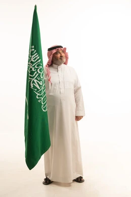 Pride and honor in belonging to the Kingdom, a portrait of an elderly Arab Saudi Gulf man smiling, wearing a ghutrah and traditional thobe, holding the Saudi flag in his hand, Flag Day March 11, celebrating the anniversary of Saudi National Day on the 8th of Rabi' al-Awwal, full-body shot, white background.