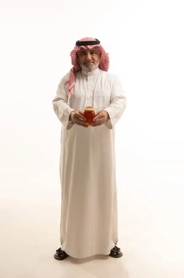 النظر إلى الكاميرا بايماءات السرور والسعادة، بورتريه مقرب لشاب عربي خليجي سعودي مسن يرتدي الثوب التقليدي يحمل بيده عبوة زجاجية من عسل النحل الطازج، منتجات وطنية محلية سعودية، غذاء طبيعي صحي، الجسم بالطول الكامل، خلفية بيضاء