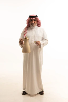 Honoring the guest and good hospitality and reception, a portrait of an elderly Arab Saudi Gulf man wearing the traditional Saudi thobe and shemagh holding in his hand the Arabic coffee pot, a local Saudi national product, gestures of cheerfulness and welcoming guests, happy Islamic holidays and occasions, full body length, white background.
