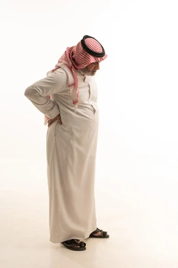 آلام العمود الفقري والظهر، بورتريه لرجل مسن عربي خليجي سعودي مبتسم يرتدي الشماغ والثوب التقليدي يضع يده على ظهره بتعابير الانحناء، إيماءات الوجع والألم، التعب والإرهاق والإحساس بالمرض، الحاجة إلى العلاج وزيارة الطبيب، صورة الجسم بالكامل، خلفية بيضاء
