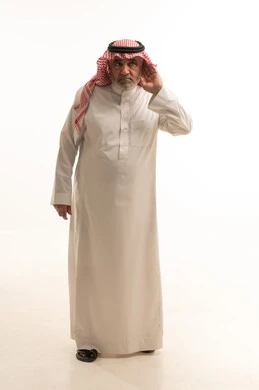 الانصات وحسن الاستماع، بورتريه لرجل مسن عربي خليجي سعودي يرتدي الثوب التقليدي والشماغ يضع يده خلف أذنه بإيماءات التنصت والتجسس، التركيز على الكلام وسماع الاسرار، صوره الجسم بالطول الكامل، خلفية بيضاء