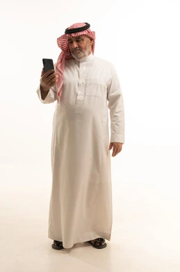 A modern and advanced technical device, elegance and handsomeness and attention to appearance, a portrait of an elderly Saudi Gulf Arab man wearing traditional attire using a mobile phone, full-body, white background.