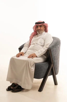 النظر إلى الكاميرا بإيماءات الجدية والثقة بالنفس، الأناقة والوسامة والاهتمام بالمظهر الخارجي، بورتريه مقرب لرجل عربي خليجي سعودي كبير السن يرتدي الزي التقليدي يجلس على كرسي مريح ، خلفية بيضاء