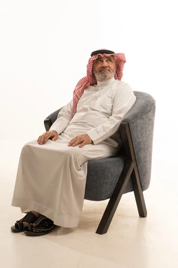 Elegance, handsomeness, and attention to appearance, gestures of seriousness and self-confidence, a close-up portrait of an elderly Saudi Arabian Gulf man wearing traditional attire sitting on a comfortable chair, with a white background.