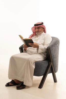 Spending free time reading, elegance and handsome appearance, a portrait of an elderly Saudi Arabian Gulf man wearing traditional attire sitting on a comfortable chair reading a book, white background.