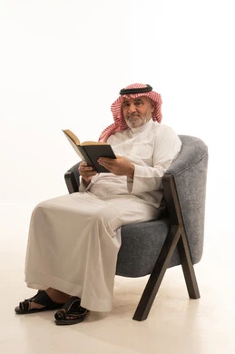 قضاء وقت الفراغ بالمطالعة، الأناقة والوسامة والاهتمام بالمظهر الخارجي، بورتريه لرجل عربي خليجي سعودي كبير السن يرتدي الزي التقليدي يجلس على كرسي مريح يقوم بقراءة كتاب، خلفية بيضاء