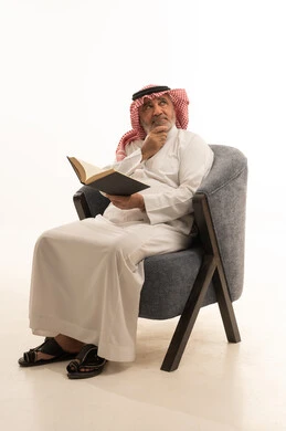 Thinking about something, spending free time reading, elegance and handsomeness and attention to appearance, a portrait of an elderly Saudi Arabian Gulf man wearing traditional attire sitting on a comfortable chair reading a book, white background.