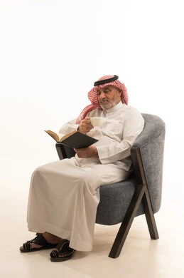 Drinking a cup of hot coffee, spending free time reading, elegance and handsomeness and attention to appearance, a portrait of an elderly Saudi Arabian Gulf man wearing traditional attire sitting on a comfortable chair reading a book, white background.