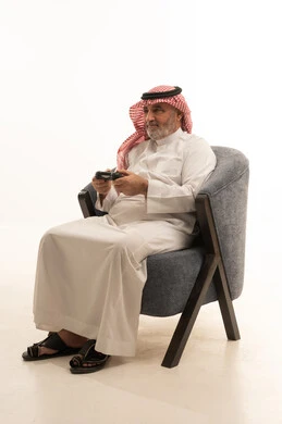 اللعب بألعاب الفيديو، التسلية وقضاء وقت الفراغ، الأناقة والوسامة والاهتمام بالمظهر الخارجي، بورتريه لرجل عربي خليجي سعودي كبير السن يرتدي الزي التقليدي يجلس على كرسي مريح يحمل بيده عصا التحكم، خلفية بيضاء