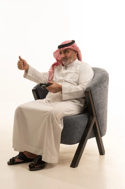 رفع اصبع الابهام بإيماءات الاعجاب، اللعب بألعاب الفيديو، التسلية وقضاء وقت الفراغ، الأناقة والوسامة والاهتمام بالمظهر الخارجي، بورتريه لرجل عربي خليجي سعودي كبير السن يرتدي الزي التقليدي يجلس على كرسي مريح يحمل بيده عصا التحكم، خلفية بيضاء