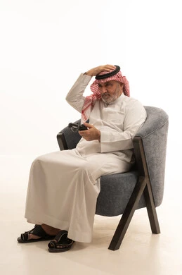 Playing video games, entertainment and spending free time, elegance and handsomeness and attention to appearance, a portrait of an elderly Arab Gulf Saudi man wearing traditional attire sitting on a comfortable chair holding a game controller, white background.
