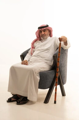 The need to use a cane, elegance and handsomeness and attention to external appearance, gestures of seriousness and self-confidence, a close-up portrait of an elderly Saudi Gulf Arab man wearing traditional attire sitting on a comfortable chair holding a cane in his hand, white background.