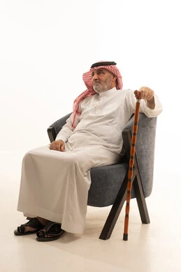The need to use a cane, elegance and handsomeness and attention to external appearance, gestures of seriousness and self-confidence, a close-up portrait of an elderly Saudi Gulf Arab man wearing traditional attire sitting on a comfortable chair holding a cane in his hand, white background.
