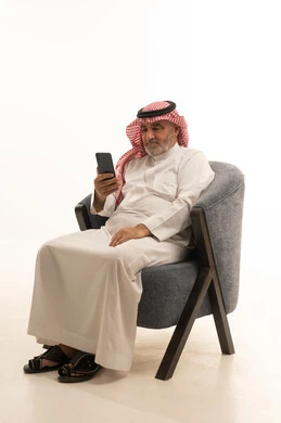 Browsing social media, communicating with family and friends, using a modern and advanced technological device, elegance and handsome appearance, a portrait of an elderly Saudi Arabian Gulf man wearing traditional attire sitting on a comfortable chair using a mobile phone, white background.