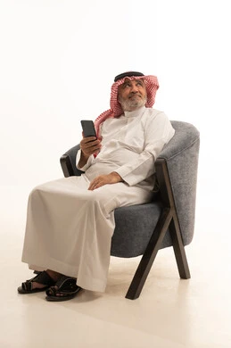 Browsing social media, communicating with family and friends, using a modern and advanced technological device, elegance and handsome appearance, a portrait of an elderly Saudi Arabian Gulf man wearing traditional attire sitting on a comfortable chair using a mobile phone, white background.