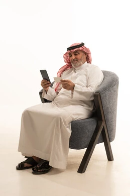 تصفح مواقع التواصل الاجتماعي، التواصل مع الاهل والاصدقاء، استخدام وسيلة تقنية حديثة ومتطورة، الأناقة والوسامة والاهتمام بالمظهر الخارجي، بورتريه لرجل عربي خليجي سعودي كبير السن يرتدي الزي التقليدي يجلس على كرسي مريح يقوم باستخدام الهاتف المحمول، خلفية بيضاء