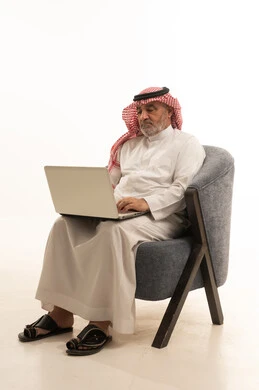 التواصل مع الاهل والاصدقاء، استخدام وسيلة تقنية حديثة ومتطورة، الأناقة والوسامة والاهتمام بالمظهر الخارجي، بورتريه لرجل عربي خليجي سعودي كبير السن يرتدي الزي التقليدي يجلس على كرسي مريح يقوم باستخدام الحاسب المحمول، خلفية بيضاء