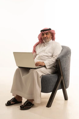 Communication with family and friends, using a modern and advanced technological means, elegance and handsomeness, and attention to external appearance, a portrait of an elderly Saudi Arabian Gulf man wearing traditional attire sitting on a comfortable chair using a laptop, white background.