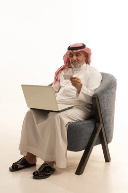 اجراء مكالمة فيديو، التواصل مع الاهل والاصدقاء، استخدام وسيلة تقنية حديثة ومتطورة، الأناقة والوسامة والاهتمام بالمظهر الخارجي، بورتريه لرجل عربي خليجي سعودي كبير السن يرتدي الزي التقليدي يجلس على كرسي مريح يقوم باستخدام الحاسب المحمول، خلفية بيضاء