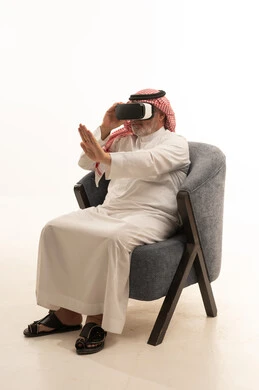 Interaction with holographic elements, elegance, handsomeness, and attention to appearance, a portrait of an elderly Saudi Arabian Gulf man wearing traditional attire sitting on a comfortable chair using virtual reality glasses, white background.
