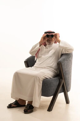 الفضول وحب الاستكشاف، رؤية الاشياء البعيدة، الأناقة والوسامة والاهتمام بالمظهر الخارجي، بورتريه لرجل عربي خليجي سعودي كبير السن يرتدي الزي التقليدي يجلس على كرسي مريح يقوم باستخدام المنظار ثنائي العينين، خلفية بيضاء