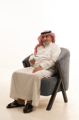 Looking at the camera with gestures of seriousness and self-confidence, elegance and handsomeness, and attention to appearance, a close-up portrait of an elderly Saudi Arabian Gulf man wearing traditional attire sitting on a comfortable chair, with a white background.