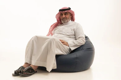 النظر الى الكاميرا بإيماءات الجدية والثقة بالنفس، الأناقة والوسامة والاهتمام بالمظهر الخارجي، بورتريه مقرب لرجل عربي خليجي سعودي كبير السن يرتدي الزي التقليدي يجلس على كرسي بين بادج مريح ، خلفية بيضاء