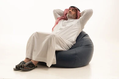 Thinking about something, the need for sleep and taking a break, elegance and handsomeness and attention to appearance, a portrait of an elderly Saudi Arabian Gulf man wearing traditional attire sitting on a comfortable chair with his hands behind his head, white background.