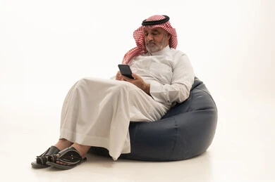 Browsing the internet, communicating with family and friends, using a modern and advanced technological device, elegance and handsomeness and attention to appearance, a portrait of an elderly Saudi Arabian Gulf man wearing traditional attire sitting on a comfortable chair using a mobile phone, white background.