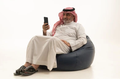 Browsing the internet, communicating with family and friends, using a modern and advanced technological device, elegance and handsomeness and attention to appearance, a portrait of an elderly Saudi Arabian Gulf man wearing traditional attire sitting on a comfortable chair using a mobile phone, white background.