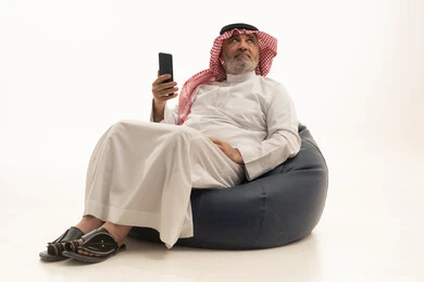 تصفح مواقع الانترنت، التواصل مع الاهل والاصدقاء، استخدام وسيلة تقنية حديثة ومتطورة، الأناقة والوسامة والاهتمام بالمظهر الخارجي، بورتريه لرجل عربي خليجي سعودي كبير السن يرتدي الزي التقليدي يجلس على كرسي مريح يقوم باستخدام الهاتف المحمول، خلفية بيضاء