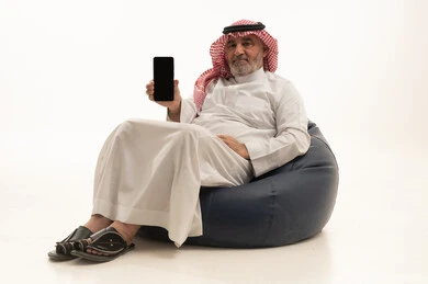 A display of a blank black screen, communication with family and friends, using a modern and advanced technological means, elegance and handsomeness and attention to external appearance, a portrait of an elderly Saudi Arabian Gulf man wearing traditional attire sitting on a comfortable chair using a mobile phone, white background.