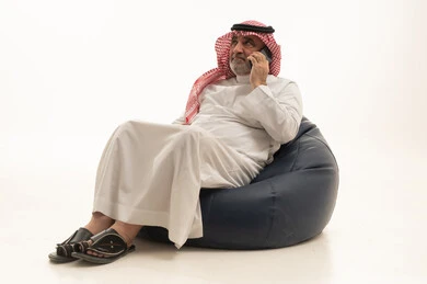 Making a phone call, communicating with family and friends, using a modern and advanced technological means, elegance and handsome appearance and attention to external appearance, a portrait of an elderly Saudi Arabian Gulf man wearing traditional attire sitting on a comfortable chair using a mobile phone, white background.