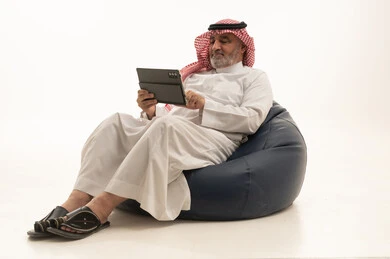 Spending time with family and friends, using a modern and advanced technological device, elegance and handsomeness, and attention to external appearance, a portrait of an elderly Arab Gulf Saudi man wearing traditional attire sitting on a comfortable chair using a mobile tablet, white background.