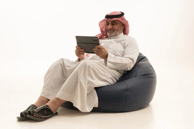 Spending time with family and friends, using a modern and advanced technological device, elegance and handsomeness, and attention to external appearance, a portrait of an elderly Arab Gulf Saudi man wearing traditional attire sitting on a comfortable chair using a mobile tablet, white background.