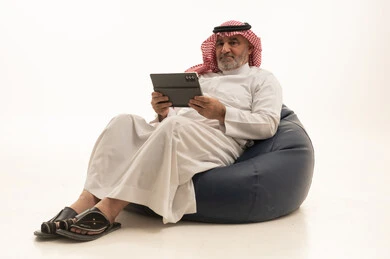 التوال مع الاهل والاصدقاء، استخدام وسيلة تقنية حديثة ومتطورة، الأناقة والوسامة والاهتمام بالمظهر الخارجي، بورتريه لرجل عربي خليجي سعودي كبير السن يرتدي الزي التقليدي يجلس على كرسي مريح يقوم باستخدام التابليت المحمول، خلفية بيضاء