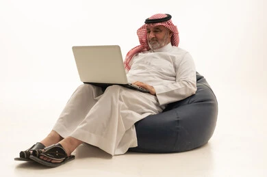 The use of a modern and advanced technological device, elegance, handsomeness, and attention to external appearance, a portrait of an elderly Saudi Arabian Gulf man wearing traditional attire sitting on a comfortable chair using a laptop, white background.