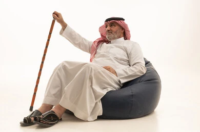 إيماءات الجدية والثقة بالنفس، الأناقة والوسامة والاهتمام بالمظهر الخارجي، بورتريه لرجل عربي خليجي سعودي كبير السن يرتدي الزي التقليدي يجلس على كرسي بين بادج مريح ويمسك بيده عكاز، خلفية بيضاء