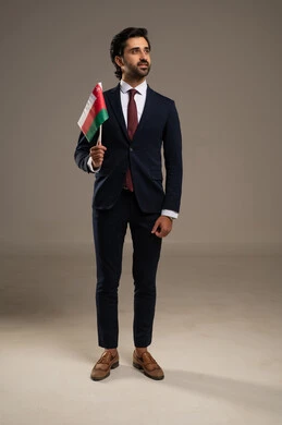 المشاركة بالاعياد والمناسبات الوطنية، الاحتفال بذكرى اليوم الوطني العماني، الاعتزاز والفخر بالرموز والراية العمانية، بورتريه لشاب عربي خليجي عماني يرتدي بدلة رسمية يحمل بيده علم سلطنة عمان، مفهوم حب الوطن والإنتماء إليه، الجسم بالطول الكامل، خلفية بيضاء